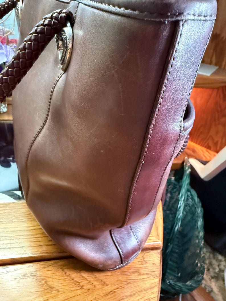 May include: A dark brown leather tote bag featuring braided leather handles attached with silver-toned metal hardware. The bag has visible vertical stitching along the side panel and a smooth, matte finish on the exterior leather surface.