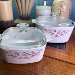 May include: Two vintage pink casserole dishes with clear glass lids. Each dish features a floral tulip design in pink and red with green leaves. The dishes are on a wooden table, with other decorative items in the background.