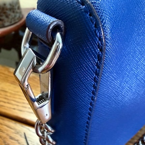 K&ouml;nnte beinhalten: Eine leuchtend blaue Handtasche mit strukturierter Kreuzschraffur-Oberfl&auml;che. Die Tasche ist mit einem gl&auml;nzenden silberfarbenen Karabinerhaken aus Metall ausgestattet, der an einer passenden Kette befestigt und seitlich gesichert ist.