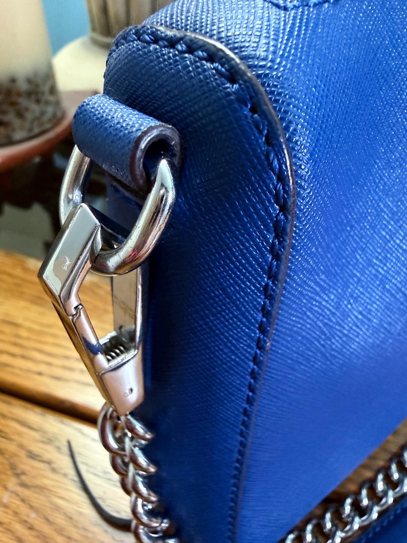 K&ouml;nnte beinhalten: Eine leuchtend blaue Handtasche mit strukturierter Kreuzschraffur-Oberfl&auml;che. Die Tasche ist mit einem gl&auml;nzenden silberfarbenen Karabinerhaken aus Metall ausgestattet, der an einer passenden Kette befestigt und seitlich gesichert ist.