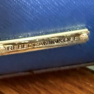 K&ouml;nnte beinhalten: Eine leuchtend blaue, texturierte Oberfl&auml;che mit einer rechteckigen, goldfarbenen Metallplakette. Die Plakette ist mit dem Text REBECCAMINKOFF in Gro&szlig;buchstaben graviert und mit kleinen Metallnieten an beiden Enden am blauen Material befestigt.