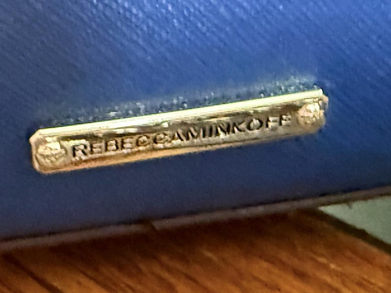 K&ouml;nnte beinhalten: Eine leuchtend blaue, texturierte Oberfl&auml;che mit einer rechteckigen, goldfarbenen Metallplakette. Die Plakette ist mit dem Text REBECCAMINKOFF in Gro&szlig;buchstaben graviert und mit kleinen Metallnieten an beiden Enden am blauen Material befestigt.