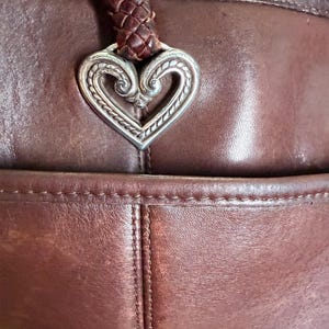 May include: A dark brown leather handbag featuring a braided leather strap attached to a silver-toned metal heart-shaped ornament with intricate embossed scrollwork details. The bag has visible stitching along the seams and a front pocket compartment.