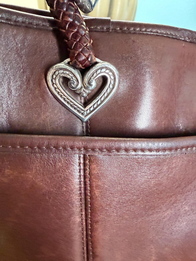 May include: A dark brown leather handbag featuring a braided leather strap attached to a silver-toned metal heart-shaped ornament with intricate embossed scrollwork details. The bag has visible stitching along the seams and a front pocket compartment.