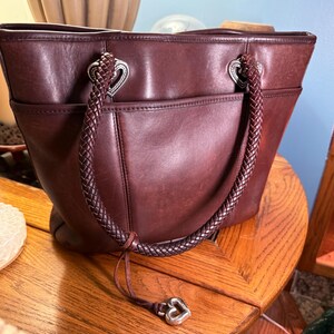 May include: A dark brown leather tote bag featuring two braided leather handles attached with silver-toned heart-shaped hardware. The bag includes an exterior slip pocket and a dangling silver-toned heart charm attached to one of the braided handles.