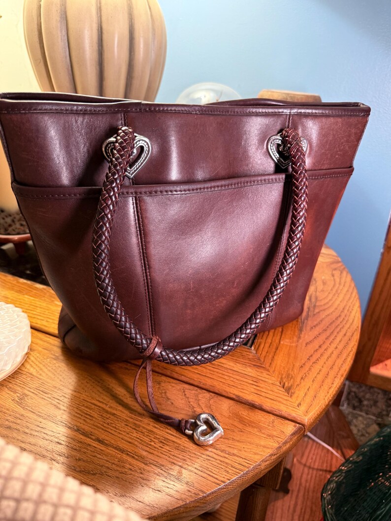 May include: A dark brown leather tote bag featuring two braided leather handles attached with silver-toned heart-shaped hardware. The bag includes an exterior slip pocket and a dangling silver-toned heart charm attached to one of the braided handles.
