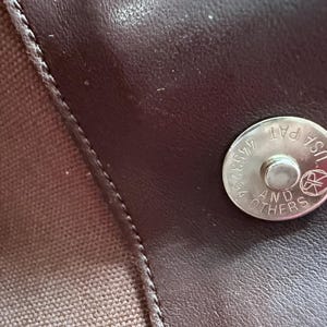 May include: A dark brown leather surface featuring a circular silver-toned magnetic snap closure. The metal fastener is engraved with the text USA PAT 4453294 AND OTHERS along with a circular logo. A textured brown fabric panel is visible on the left.