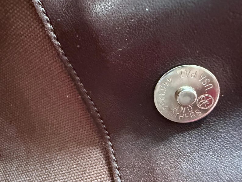May include: A dark brown leather surface featuring a circular silver-toned magnetic snap closure. The metal fastener is engraved with the text USA PAT 4453294 AND OTHERS along with a circular logo. A textured brown fabric panel is visible on the left.