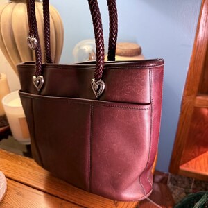 May include: A deep burgundy leather tote bag featuring two braided dark brown leather handles. The handles are attached to the bag with silver-toned metal heart-shaped hardware. The bag has a horizontal seam across the front and a slip pocket.