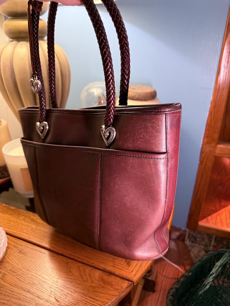 May include: A deep burgundy leather tote bag featuring two braided dark brown leather handles. The handles are attached to the bag with silver-toned metal heart-shaped hardware. The bag has a horizontal seam across the front and a slip pocket.
