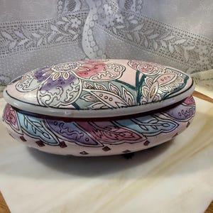 May include: Oval-shaped trinket box with a lid, featuring a pastel floral design in pink, purple, and blue. The box has a light pink base with a dark red interior and a decorative border. The floral pattern includes intricate leaf and petal details.