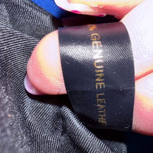 K&ouml;nnte beinhalten: Ein schwarzes Stoffetikett mit goldenem Text, auf dem 100% GENUINE LEATHER steht. Das Etikett ist an einem schwarzen, strukturierten Innenfutter befestigt. Der Text ist deutlich lesbar und vertikal auf dem rechteckigen, glatten Gewebeetikett angebracht.