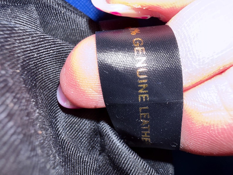 K&ouml;nnte beinhalten: Ein schwarzes Stoffetikett mit goldenem Text, auf dem 100% GENUINE LEATHER steht. Das Etikett ist an einem schwarzen, strukturierten Innenfutter befestigt. Der Text ist deutlich lesbar und vertikal auf dem rechteckigen, glatten Gewebeetikett angebracht.