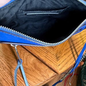 K&ouml;nnte beinhalten: Eine leuchtend blaue Umh&auml;ngetasche mit einem silberfarbenen Metallrei&szlig;verschluss. Das Innere ist mit schwarzem Stoff ausgekleidet und verf&uuml;gt &uuml;ber ein Einsteckfach. Ein d&uuml;nner blauer Lederanh&auml;nger ist am Rei&szlig;verschluss befestigt, und ein blauer Riemen ist enthalten.