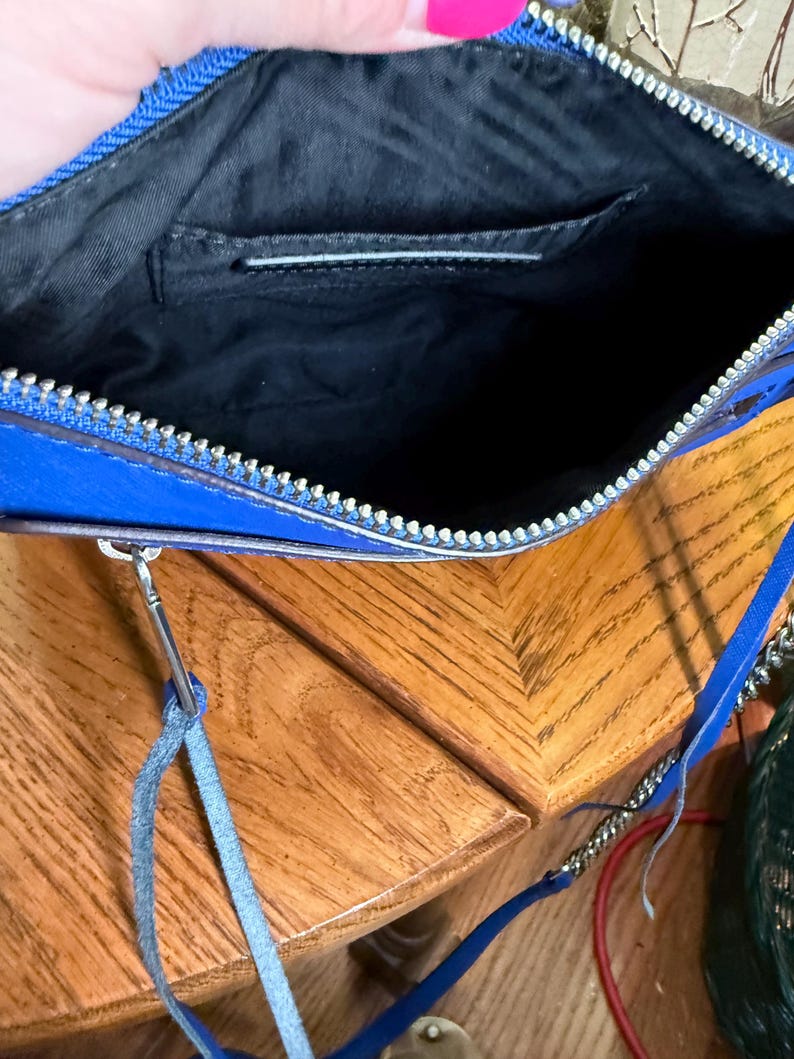 K&ouml;nnte beinhalten: Eine leuchtend blaue Umh&auml;ngetasche mit einem silberfarbenen Metallrei&szlig;verschluss. Das Innere ist mit schwarzem Stoff ausgekleidet und verf&uuml;gt &uuml;ber ein Einsteckfach. Ein d&uuml;nner blauer Lederanh&auml;nger ist am Rei&szlig;verschluss befestigt, und ein blauer Riemen ist enthalten.