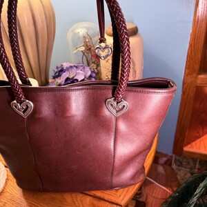 May include: A dark brown leather tote bag featuring two braided leather handles attached with silver-toned heart-shaped metal hardware. A small silver heart-shaped charm hangs from one of the handles, and the bag has a smooth, matte finish.