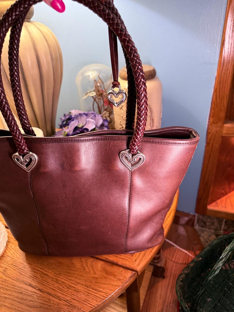 May include: A dark brown leather tote bag featuring two braided leather handles attached with silver-toned heart-shaped metal hardware. A small silver heart-shaped charm hangs from one of the handles, and the bag has a smooth, matte finish.