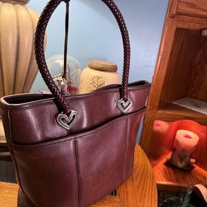 May include: A dark brown leather tote bag featuring a single braided handle attached with silver-toned heart-shaped metal hardware. The bag has a smooth finish with visible stitching details and an open exterior pocket on the front side.