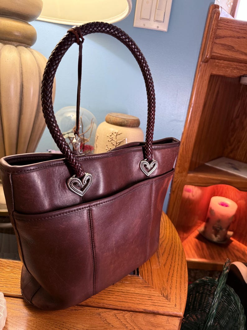 May include: A dark brown leather tote bag featuring a single braided handle attached with silver-toned heart-shaped metal hardware. The bag has a smooth finish with visible stitching details and an open exterior pocket on the front side.