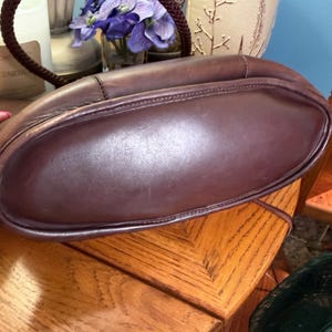 May include: A dark brown leather handbag featuring a smooth, rounded base and a braided leather handle. The bag has visible stitching along the seams and a structured, curved silhouette. The leather has a matte finish with subtle natural variations.