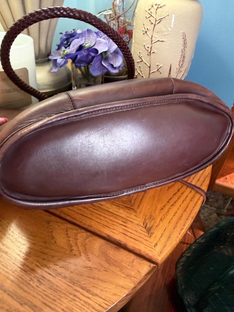 May include: A dark brown leather handbag featuring a smooth, rounded base and a braided leather handle. The bag has visible stitching along the seams and a structured, curved silhouette. The leather has a matte finish with subtle natural variations.
