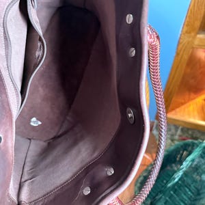 May include: A dark brown leather tote bag interior featuring a smooth fabric lining, a zippered side pocket, and a magnetic snap closure. The bag includes a braided leather handle attached with silver-toned metal rivets along the top edge.