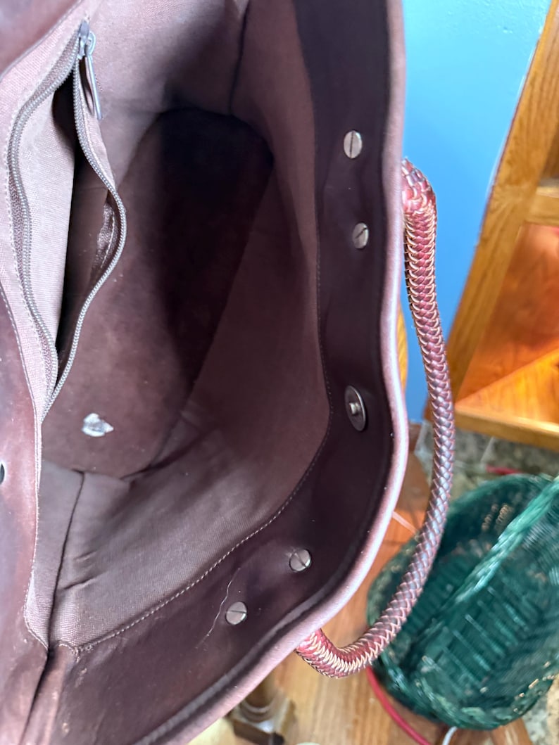 May include: A dark brown leather tote bag interior featuring a smooth fabric lining, a zippered side pocket, and a magnetic snap closure. The bag includes a braided leather handle attached with silver-toned metal rivets along the top edge.