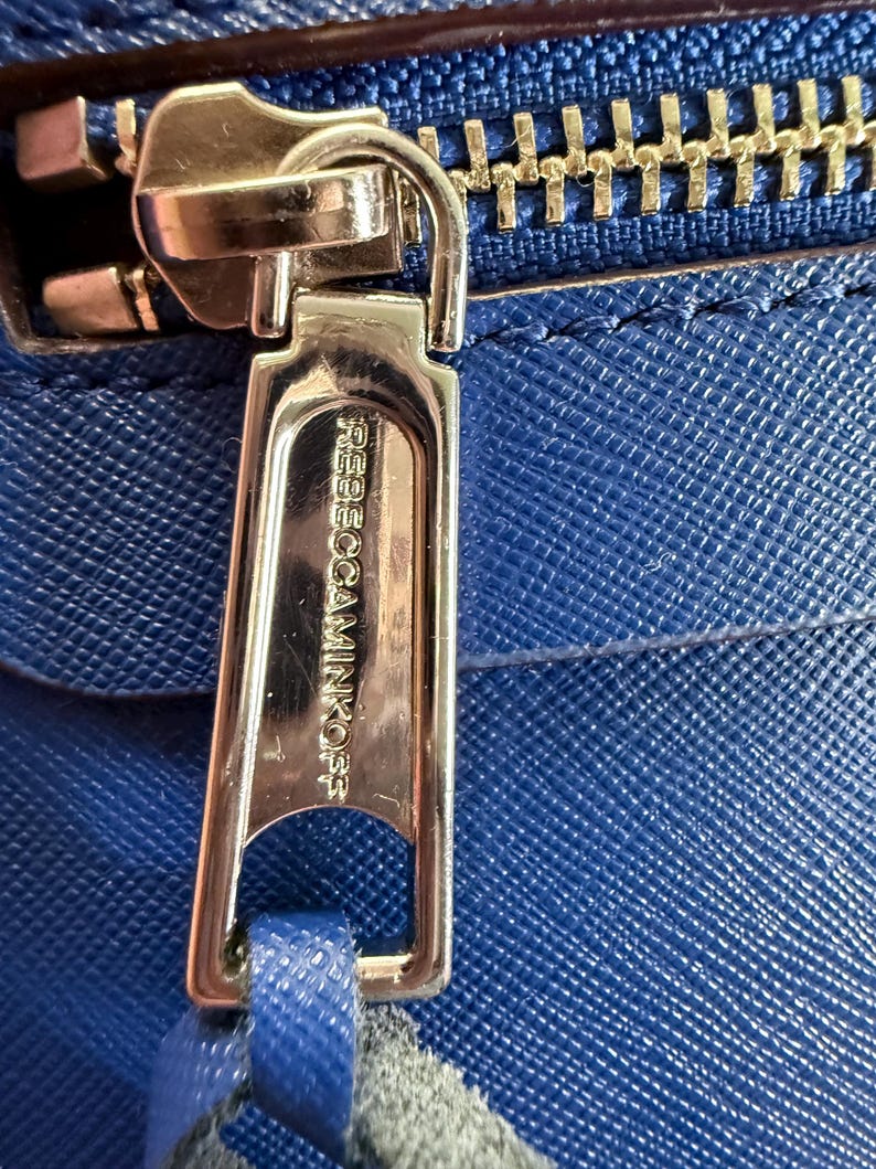K&ouml;nnte beinhalten: Eine leuchtend blaue Handtasche aus strukturiertem Leder mit einem polierten goldfarbenen Rei&szlig;verschlussanh&auml;nger. Der Anh&auml;nger ist mit dem Text REBECCAMINKOFF graviert. Eine passende blaue Lederlasche ist an der Metallschlie&szlig;e befestigt.