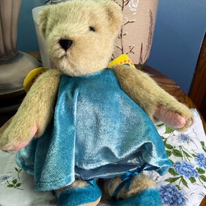 May include: A plush teddy bear wearing a turquoise blue dress and matching shoes. The bear has tan fur, a black nose, and pink accents on its paws. A yellow tag is attached to the bear's back. The bear is sitting on a floral patterned cloth.