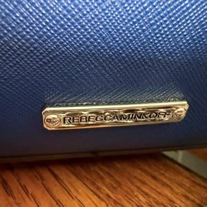 K&ouml;nnte beinhalten: Eine leuchtend k&ouml;nigsblaue Handtasche mit strukturierter Oberfl&auml;che und einer polierten, rechteckigen Metallplakette in Silberoptik. Die Plakette ist mit zwei kleinen Schrauben befestigt und zeigt den Markennamen REBECCAMINKOFF in Relief.