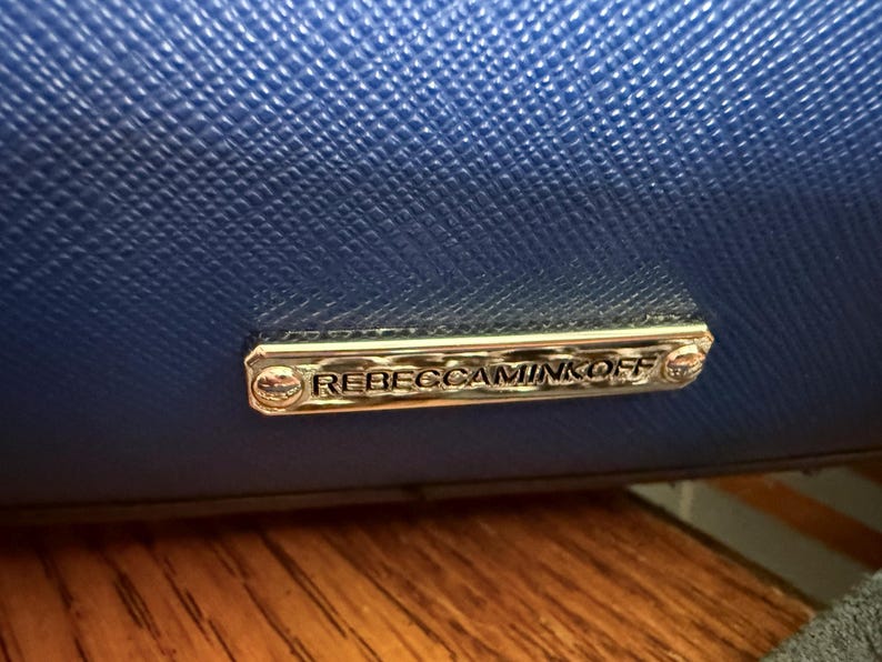 K&ouml;nnte beinhalten: Eine leuchtend k&ouml;nigsblaue Handtasche mit strukturierter Oberfl&auml;che und einer polierten, rechteckigen Metallplakette in Silberoptik. Die Plakette ist mit zwei kleinen Schrauben befestigt und zeigt den Markennamen REBECCAMINKOFF in Relief.