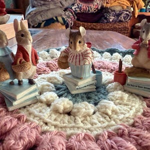 May include: A collection of figurines depicting storybook characters, including rabbits, a mouse, and a squirrel. The figurines are standing on small stacks of books, and are dressed in detailed clothing. The scene is set on a colorful, crocheted blanket.