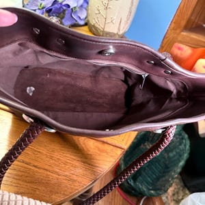 May include: A dark brown tote bag interior featuring a smooth lining, a zippered pocket along the back wall, and a magnetic snap closure at the top. The bag includes two braided dark brown handles attached with silver-toned metal hardware.