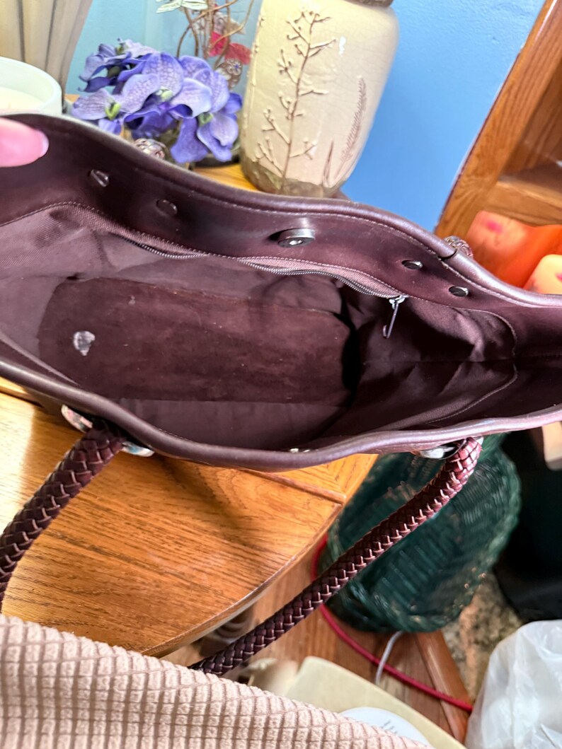 May include: A dark brown tote bag interior featuring a smooth lining, a zippered pocket along the back wall, and a magnetic snap closure at the top. The bag includes two braided dark brown handles attached with silver-toned metal hardware.