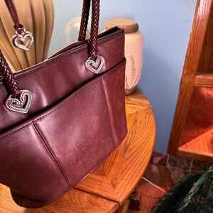 May include: A deep burgundy leather tote bag featuring braided leather handles attached with silver-toned heart-shaped metal hardware. The bag includes a front exterior pocket and a dangling silver heart charm attached to one of the handles.