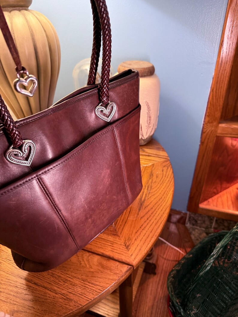 May include: A deep burgundy leather tote bag featuring braided leather handles attached with silver-toned heart-shaped metal hardware. The bag includes a front exterior pocket and a dangling silver heart charm attached to one of the handles.