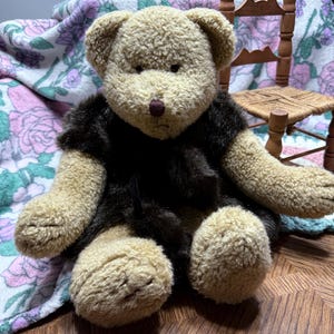May include: A plush teddy bear with tan fur and a dark brown vest. The bear has a brown nose and is sitting on a wooden surface. A floral blanket and a miniature wooden chair are in the background.