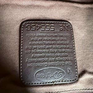 May include: A dark brown rectangular leather patch sewn onto a brown fabric lining. The patch features the embossed Brighton logo, a V symbol, and the text: The Brighton Collection, handbags are individually crafted from quality leathers. N&deg; 555456.