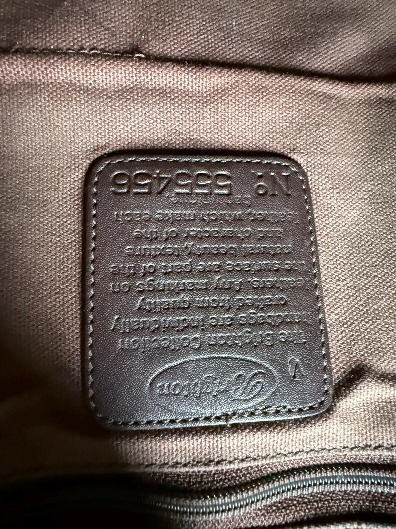 May include: A dark brown rectangular leather patch sewn onto a brown fabric lining. The patch features the embossed Brighton logo, a V symbol, and the text: The Brighton Collection, handbags are individually crafted from quality leathers. N&deg; 555456.