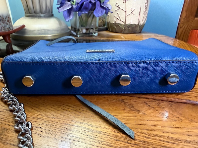 K&ouml;nnte beinhalten: Eine leuchtend blaue, rechteckige Handtasche mit strukturierter Oberfl&auml;che, ausgestattet mit vier silberfarbenen Metallf&uuml;&szlig;en an der Unterseite und einem silberfarbenen Metallkettenriemen. Eine kleine, rechteckige silberne Metallplatte ist vorne mittig.