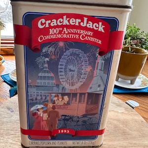 May include: A vintage Cracker Jack tin canister commemorating the 100th anniversary of the brand. The tin features a colorful illustration of a Ferris wheel and a red banner with the text "Cracker Jack 100th Anniversary Commemorative Canister". The bottom of the tin reads "Caramel Popcorn and Peanuts Net Wt. 8 oz. (226g)" and "1893".
