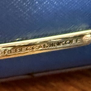 K&ouml;nnte beinhalten: Eine leuchtend blaue, texturierte Oberfl&auml;che mit einer rechteckigen, goldfarbenen Metallplakette, die am Material befestigt ist. Die Plakette ist mit dem Text REBECCAMINKOFF in Gro&szlig;buchstaben graviert und hat kleine dekorative Schraubendetails.