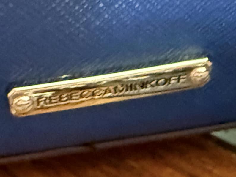 K&ouml;nnte beinhalten: Eine leuchtend blaue, texturierte Oberfl&auml;che mit einer rechteckigen, goldfarbenen Metallplakette, die am Material befestigt ist. Die Plakette ist mit dem Text REBECCAMINKOFF in Gro&szlig;buchstaben graviert und hat kleine dekorative Schraubendetails.
