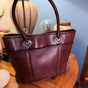 May include: A deep burgundy leather tote bag featuring a single braided leather handle. The handle attaches to the bag with silver-toned metal heart-shaped hardware. The bag has a structured silhouette with a front pocket and smooth leather finish.
