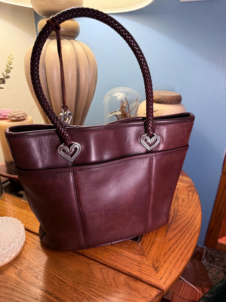 May include: A deep burgundy leather tote bag featuring a single braided leather handle. The handle attaches to the bag with silver-toned metal heart-shaped hardware. The bag has a structured silhouette with a front pocket and smooth leather finish.