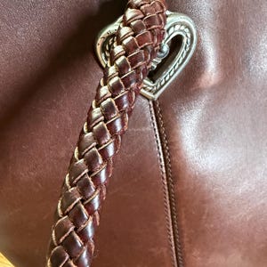 May include: A dark brown leather handbag featuring a thick, braided leather handle attached to the bag with a silver-toned metal heart-shaped hardware piece. The leather surface shows a smooth texture with visible stitching along the seams.