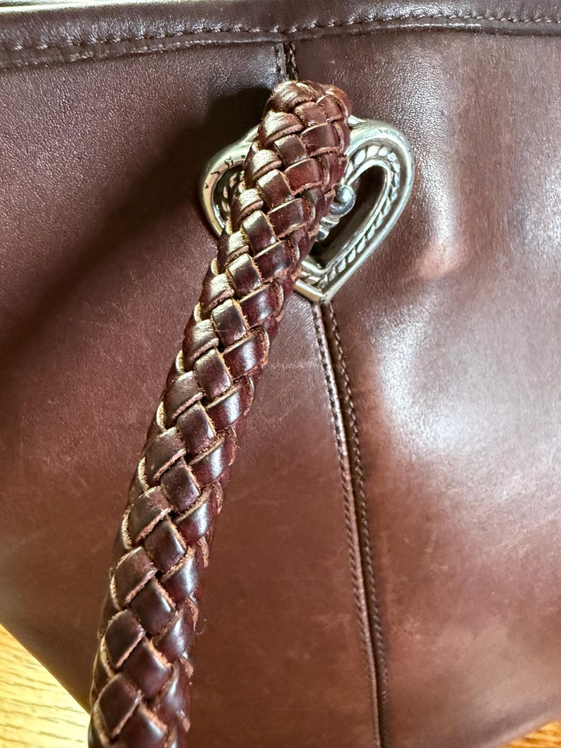 May include: A dark brown leather handbag featuring a thick, braided leather handle attached to the bag with a silver-toned metal heart-shaped hardware piece. The leather surface shows a smooth texture with visible stitching along the seams.