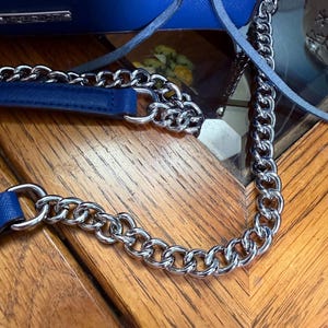 K&ouml;nnte beinhalten: Eine leuchtend blaue Handtasche mit strukturierter Oberfl&auml;che und einem silberfarbenen Metallkettengurt. Die Tasche verf&uuml;gt &uuml;ber ein passendes, in die Kette integriertes Schulterpolster aus blauem Kunstleder und eine kleine silberne Metallplatte.