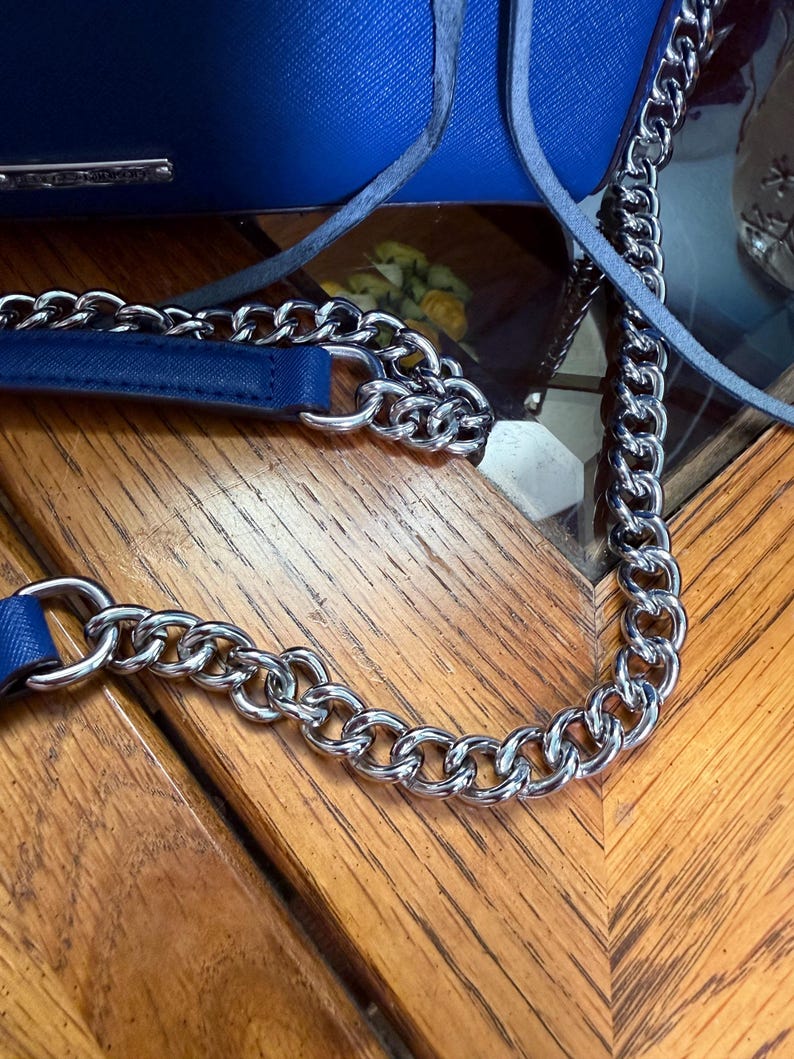 K&ouml;nnte beinhalten: Eine leuchtend blaue Handtasche mit strukturierter Oberfl&auml;che und einem silberfarbenen Metallkettengurt. Die Tasche verf&uuml;gt &uuml;ber ein passendes, in die Kette integriertes Schulterpolster aus blauem Kunstleder und eine kleine silberne Metallplatte.