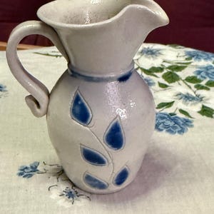 May include: A small, off-white ceramic pitcher with a curved handle and a spout. The pitcher is decorated with blue leaf-shaped designs and a blue band around the neck. The pitcher is likely handmade and has a rustic, artisanal feel.